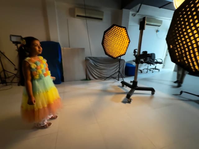 This image features a child standing in what appears to be a photography studio. The child is looking at the camera, wearing a colorful outfit, with their arms by their sides. In front of them on the floor are several items that resemble photo backdrops or props, including what seems like a honeycomb pattern and possibly other elements for photoshoots. The background is dimly lit, which suggests it might be evening or in a room without artificial lighting. The setting gives an impression of being in a professional photographic environment.