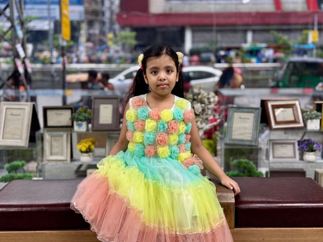The image features a young girl with a colorful dress, posing in front of a display that includes various items on shelves. She appears to be sitting at a bench and has a sweet and joyful expression. Behind her, there seems to be an indoor environment with framed pictures on the wall. The setting suggests this could be a public space like a museum or a gift shop.