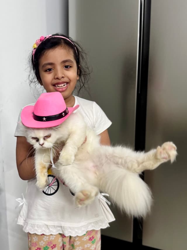 In the image, a young girl is standing next to a cat. The cat is wearing a pink hat. The girl appears to be holding the cat, who seems to be comfortable with her touch. She is dressed in casual attire and looks happy to have her pet cat with her.
