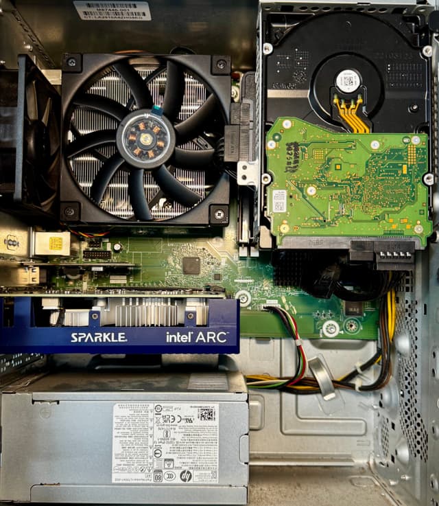 The image is an interior view of a computer's hardware. It includes a CPU socket with a processor installed, multiple RAM slots with RAM modules, a graphics card with a fan attached, a motherboard with various connectors and expansion slots visible, as well as a power supply unit and a hard drive.