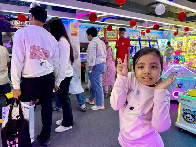 This image captures a lively indoor setting, likely an arcade or game center. In the foreground, there is a young girl with a cheerful expression, making a peace sign gesture with her fingers. She appears to be dressed in casual attire and has a playful pose. Behind her, several people are engaged in various activities; some are standing near colorful machines or games, while others are interacting with what seems to be a booth selling tickets or items. The environment is vibrant with neon lights and bright colors, contributing to the festive atmosphere. There are also balloons, signs, and other decorations visible, suggesting this could be an event or celebration.