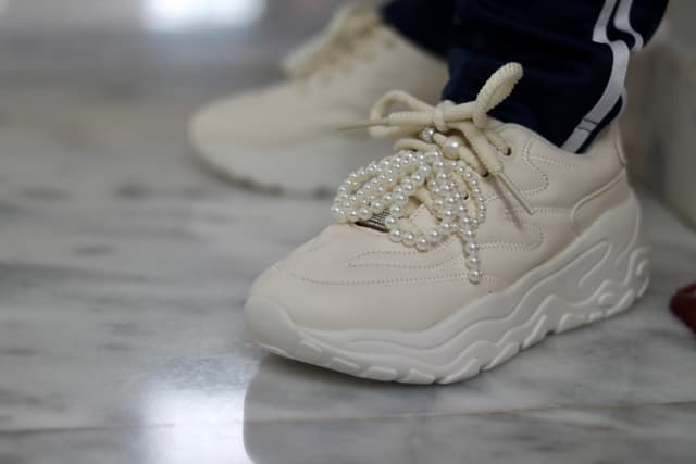 The image is a close-up photograph of a pair of white sneakers worn by someone standing on a smooth, reflective surface. The sneakers feature a lace-up design with a small, round knob at the top of each shoe, possibly for adjusting the fit. The soles appear to be chunky and flat. There are no visible texts or logos in this image. The background is minimalistic and nondescript, with a light-colored surface and a soft, blurred background that suggests a focus on the shoes themselves.