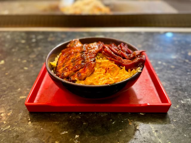 The image features an oriental meal served on a red tray atop a countertop. There are two bowls: one with what appears to be steamed rice and another with slices of grilled meat, possibly chicken. Beside the dishes are a small bowl filled with a sauce that could be chili or curry, and a portion of fried noodles in a separate bowl. The tabletop has a dark surface, and the overall setting suggests a casual dining environment. There is also a blurred image of what looks like another food item in the background, possibly part of the same meal or from a nearby station.