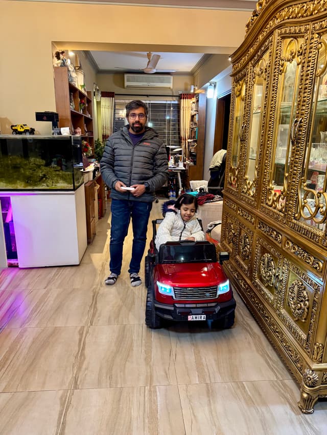 The image depicts an indoor space, possibly a showroom or a shop floor given the wooden floor and a variety of items on display. In the foreground, there is a red motorized cart with one person sitting inside it. Standing to the left of the cart is a man who appears to be waiting for something or someone; he's holding what looks like an electronic device in his hands. The shop has a variety of products displayed on shelves and tables, including what seems to be jewelry. In the background, there's another person visible from the waist up, suggesting more customers present in the store. There's no immediate text within the image itself to provide additional context.