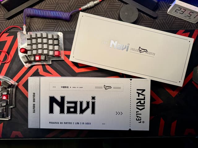 The image shows a desk with various items. There's a metal box labeled "NAVI," which appears to be an electronic device or component. Next to it, there are keyboards and what looks like a computer mouse, suggesting that this might be a workspace for someone who uses a computer. There's also a small model ship, indicating an interest in space or a collectible hobby. The overall setting suggests a room dedicated to electronics or hobbies related to technology and space exploration.