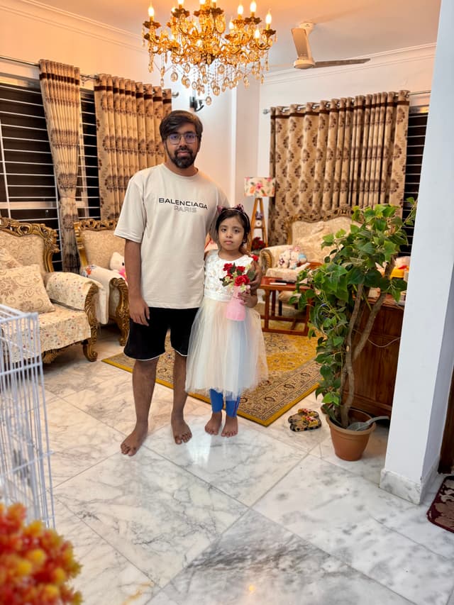 The image captures a moment inside a home. In the foreground, there's an adult and a young child standing in front of a couch. They are dressed casually and appear to be posing for the photo. Behind them, the living room is furnished with a sofa and a coffee table that has a couple of items on it. The space features a large window with curtains partially drawn, allowing natural light into the room. On the left side of the image, there's a wall-mounted air conditioning unit, and on the right, there is decorative wall art. The overall atmosphere suggests a comfortable domestic setting.