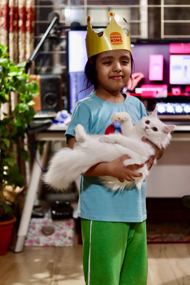 In the image, there's a child standing on what appears to be a carpeted floor in front of a TV. The child is holding a white cat that has a fluffy tail and ears. The child is wearing a t-shirt with a graphic design and shorts, along with dark pants. Behind the child, there's a table with some objects on it, and a potted plant is visible to the right. There's a hint of an adult sitting to the left in front of the TV.