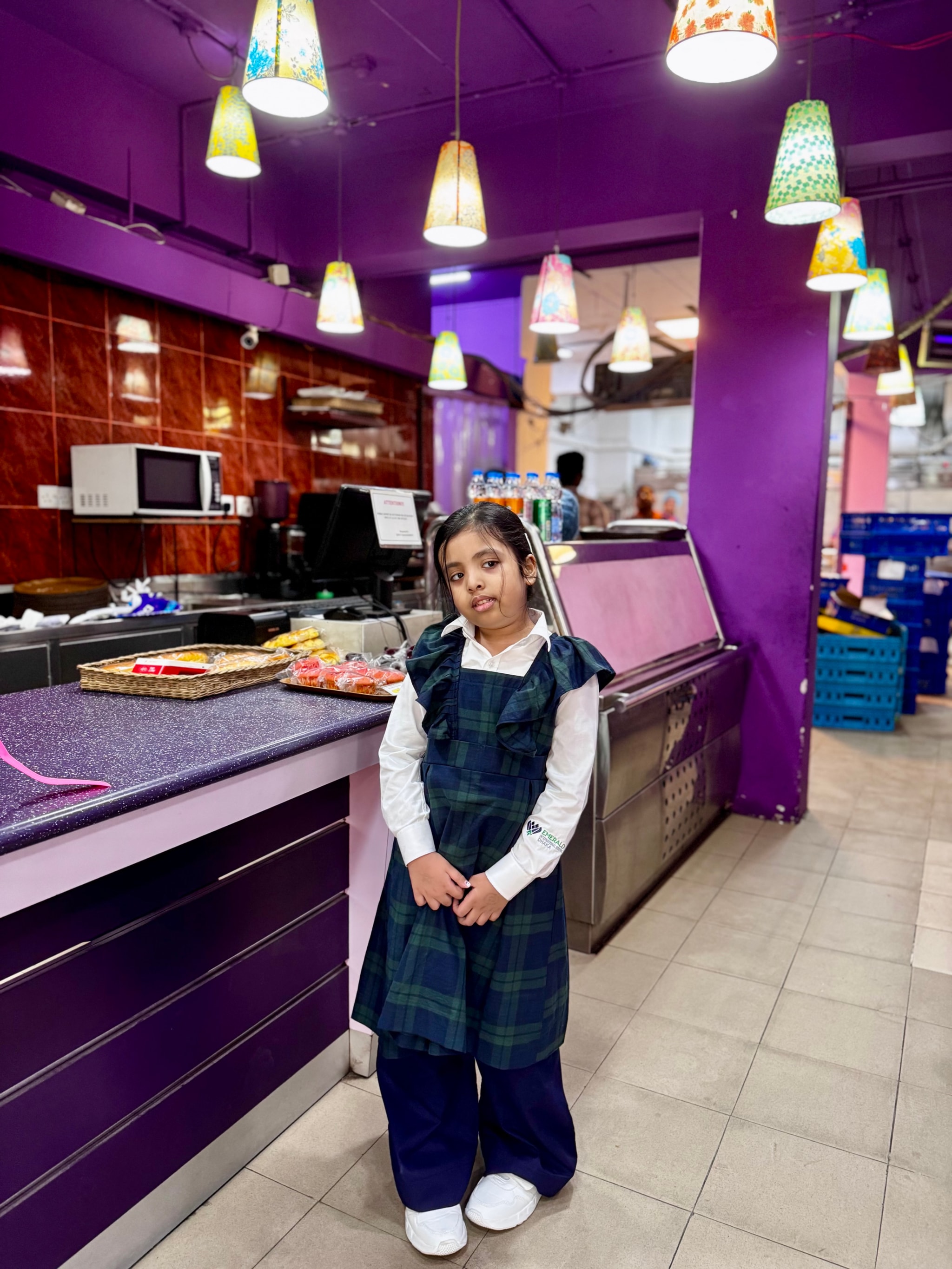 In the image, there's a young girl standing in front of a counter. The counter has a variety of pastries and cakes on display. Behind her, there are shelves with more items for sale. On either side of her, there are decorative lights hanging from the ceiling. To the right, there is a small part of what appears to be an establishment with a purple-themed interior. The girl seems to be posing for the photo with a slight smile on her face.