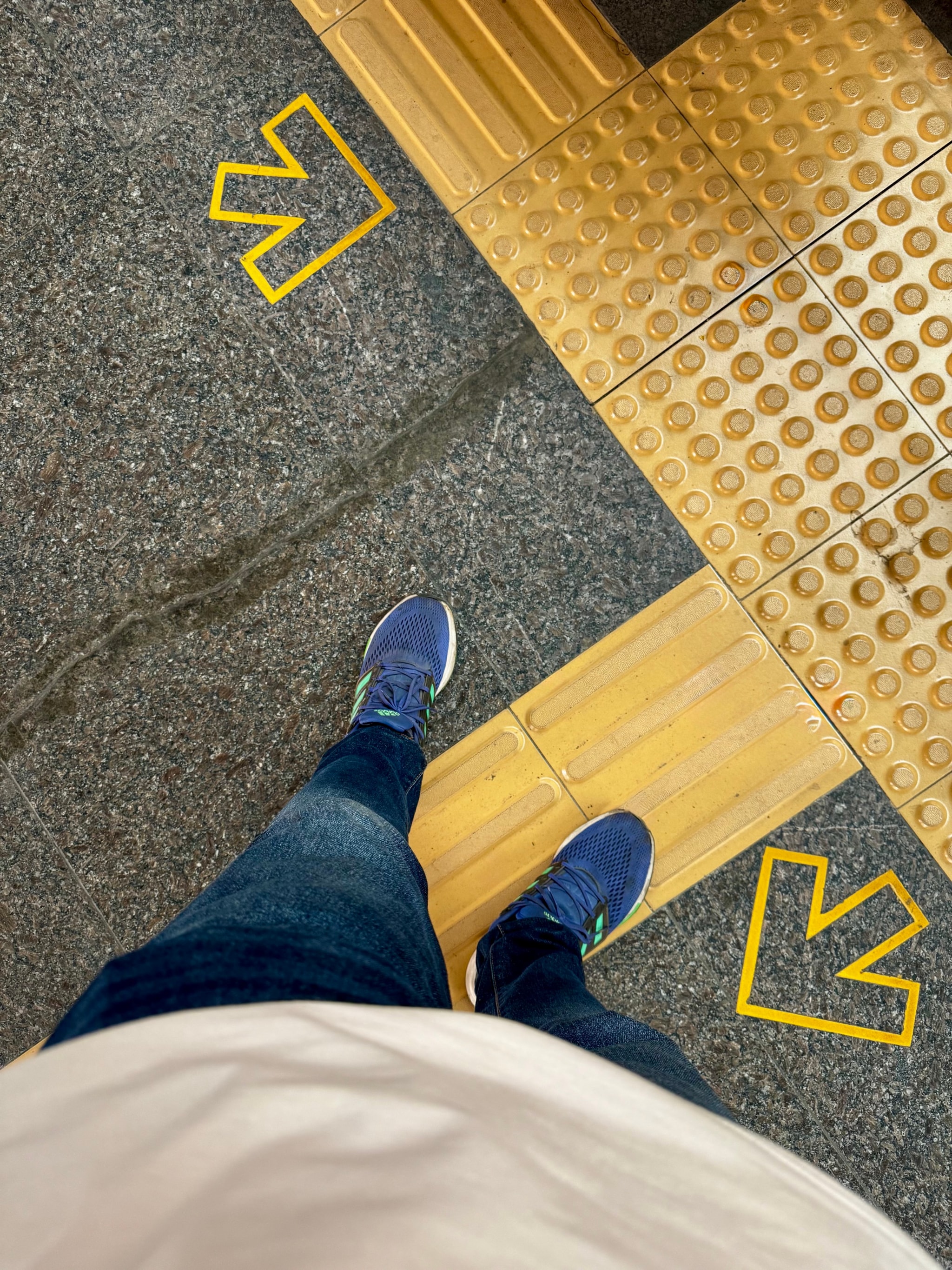 The image features a person standing on an escalator at what appears to be a subway station. The individual is wearing a white t-shirt, blue jeans, and athletic shoes with a checkmark design. They are also wearing shorts. The escalator has yellow lines painted on the right side, indicating directions for passengers. At the top of the image, there are two arrows pointing to different directions, suggesting that the person is at a junction or change of direction in the station's layout.