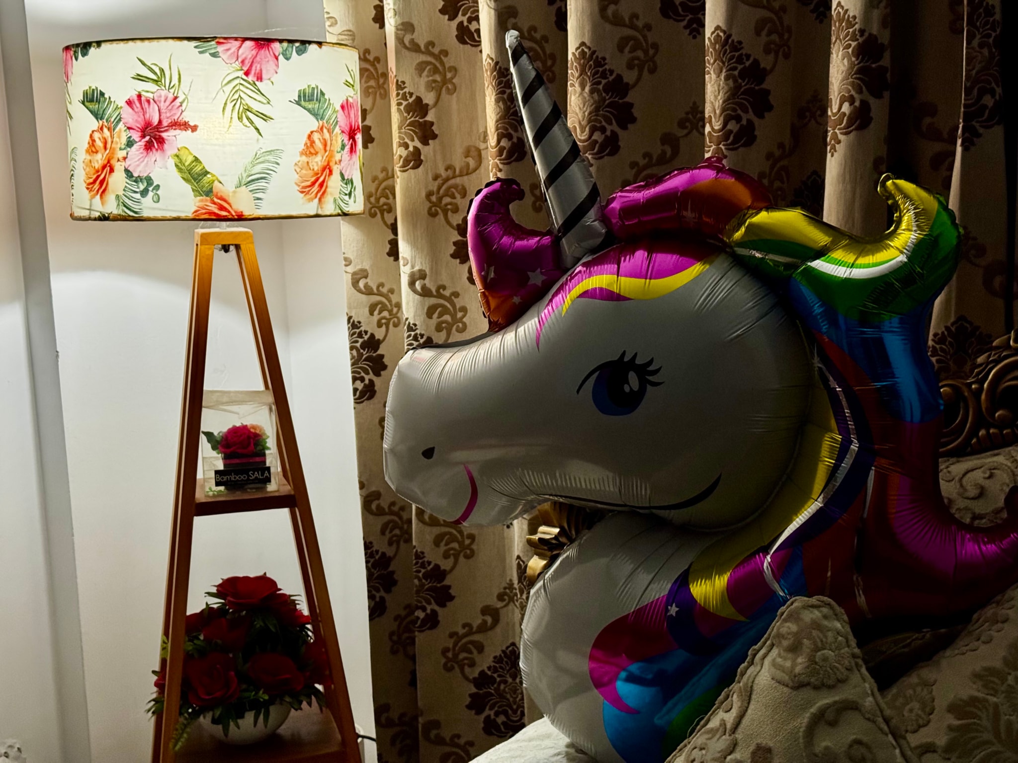 The image features a colorful toy unicorn sitting on a surface, placed next to a white wall. The unicorn has a large mane and tail, with different shades of pink and purple. It stands upright, with one leg elevated. Behind the unicorn, there is a framed picture or artwork hanging on the wall. Next to this frame is a tall wooden ladder leaning against it. To the left of the unicorn, a lamp with a floral shade casts light onto part of the unicorn's body. The background has a patterned fabric and a portion of a window can be seen.