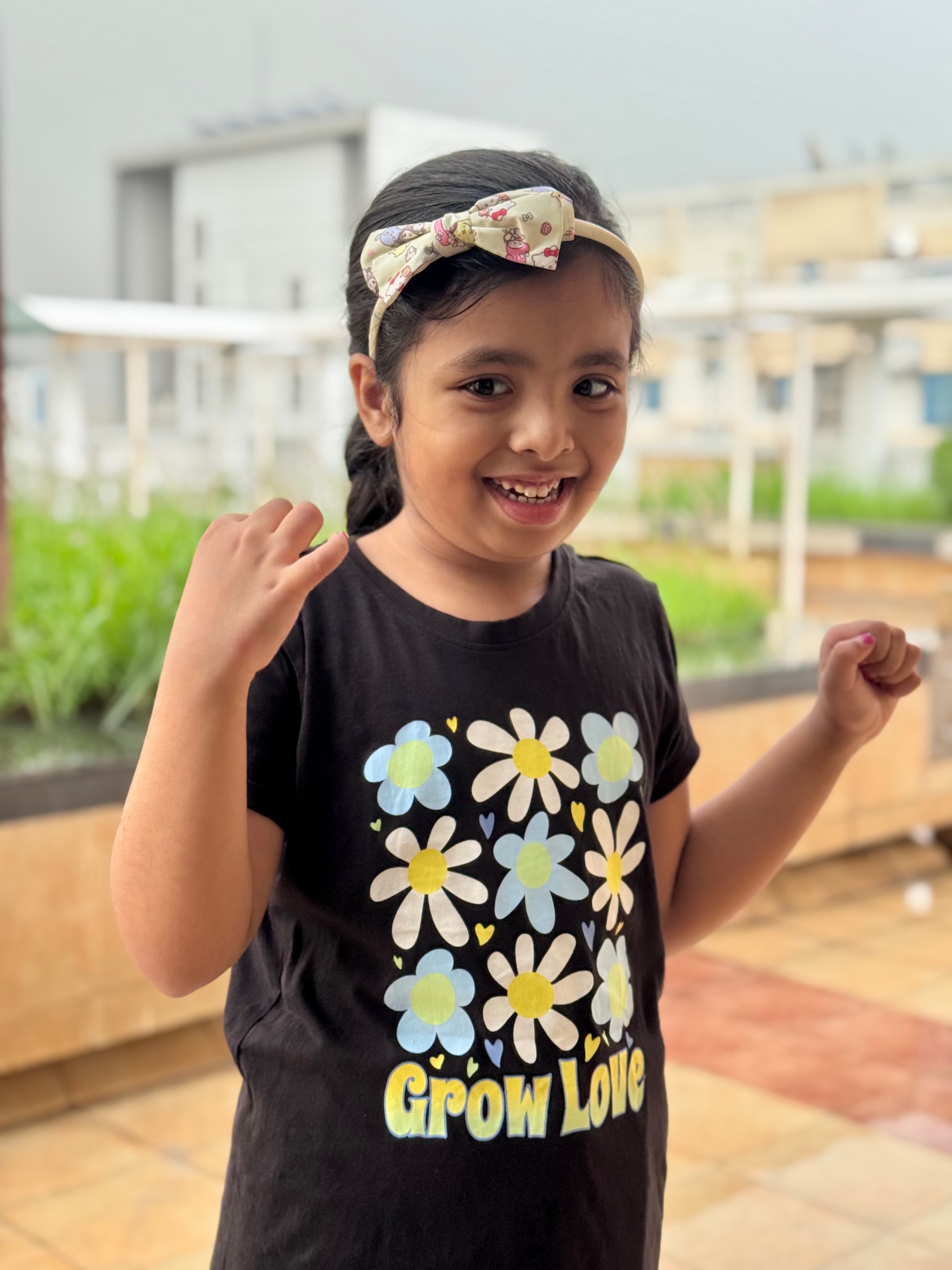 The image depicts a young girl outdoors. She has a playful expression, smiling and holding her hair with her left hand. Her right hand is raised slightly above her shoulder. She's wearing a black shirt with a floral print on the front, along with a white headband adorned with colorful flowers. The child appears to be standing in front of a building or structure with some foliage nearby. There are no visible texts within the image.