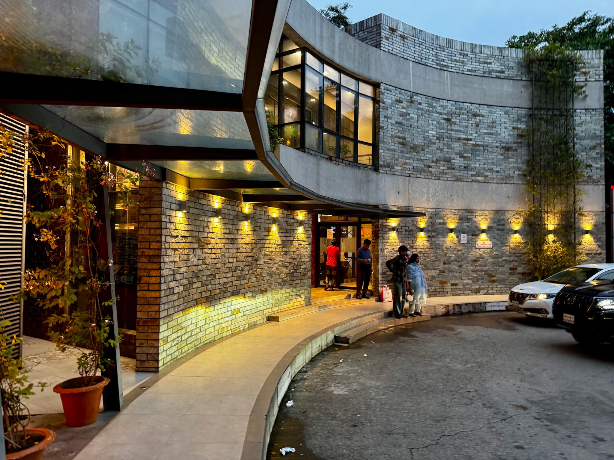The image captures an exterior view of a modern restaurant with a distinctive architectural design. The facade is characterized by curved walls and a series of recessed lighting fixtures that illuminate the exterior, enhancing its contemporary feel. A circular window punctuates one of the walls, contributing to the unique aesthetic. The restaurant appears to be open for business, as indicated by the visible interior lights. In front of the restaurant, there is a neatly maintained driveway with a small tree planted along it, adding a touch of greenery to the urban setting. A car is parked in front of the entrance, suggesting recent activity or patrons. The surrounding area is not highly detailed but seems to be an outdoor space with potted plants and other architectural elements. The image is taken during what appears to be evening or nighttime due to the artificial lighting and the darkness visible beyond the restaurant's immediate surroundings.