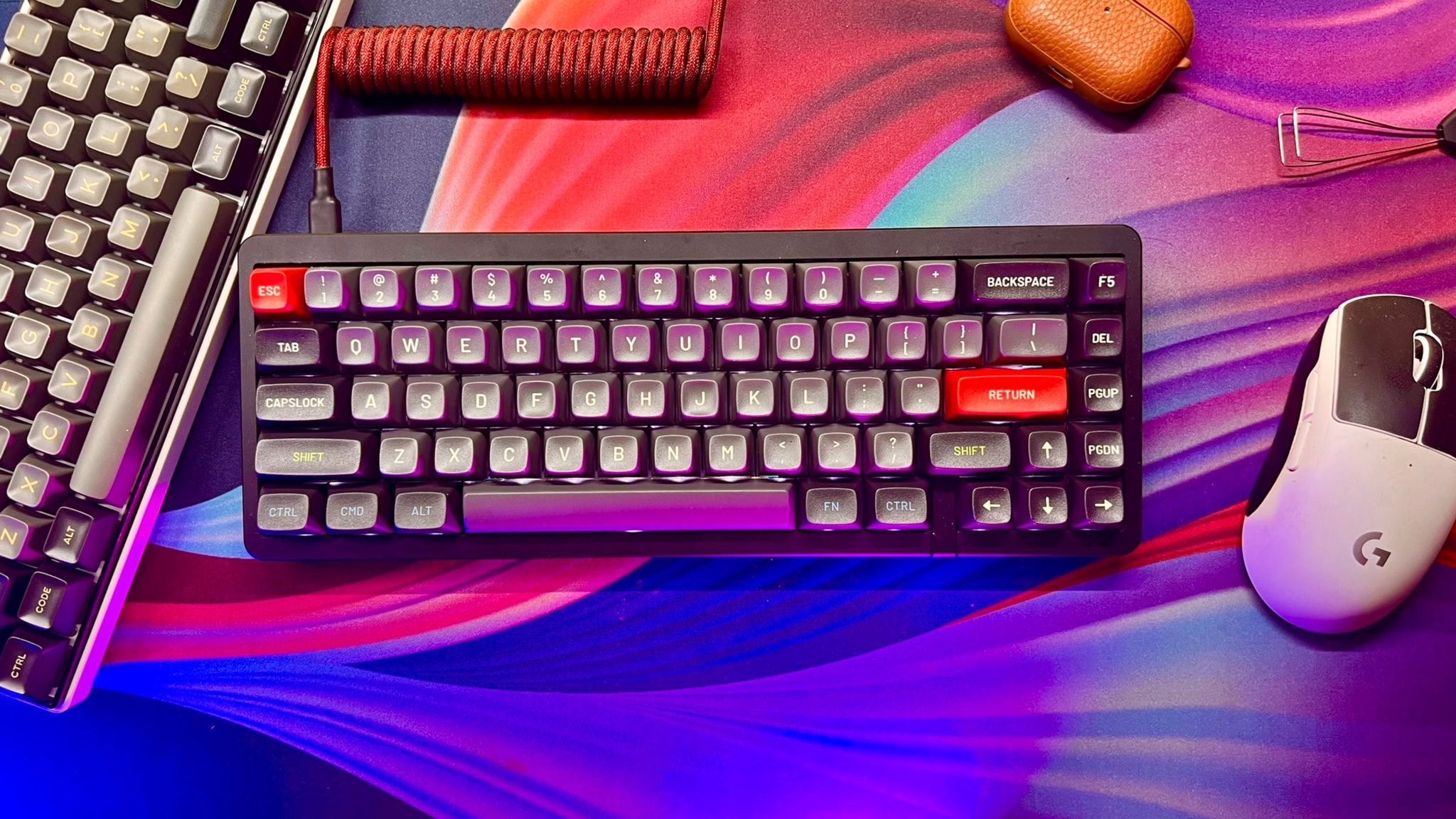 The image displays a desktop computer setup on a colorful surface with a vibrant, fluid-like design. There's a mechanical keyboard with red keycaps in the foreground, and to its right is a gaming mouse. In the background, there's a gaming keyboard with gray keys and a wrist rest. Additionally, there are a few objects that seem to be part of the setup, such as a pen holder with pens, and possibly a piece of paper or a notepad. The style of the image is a standard photograph capturing this setup from an angle.