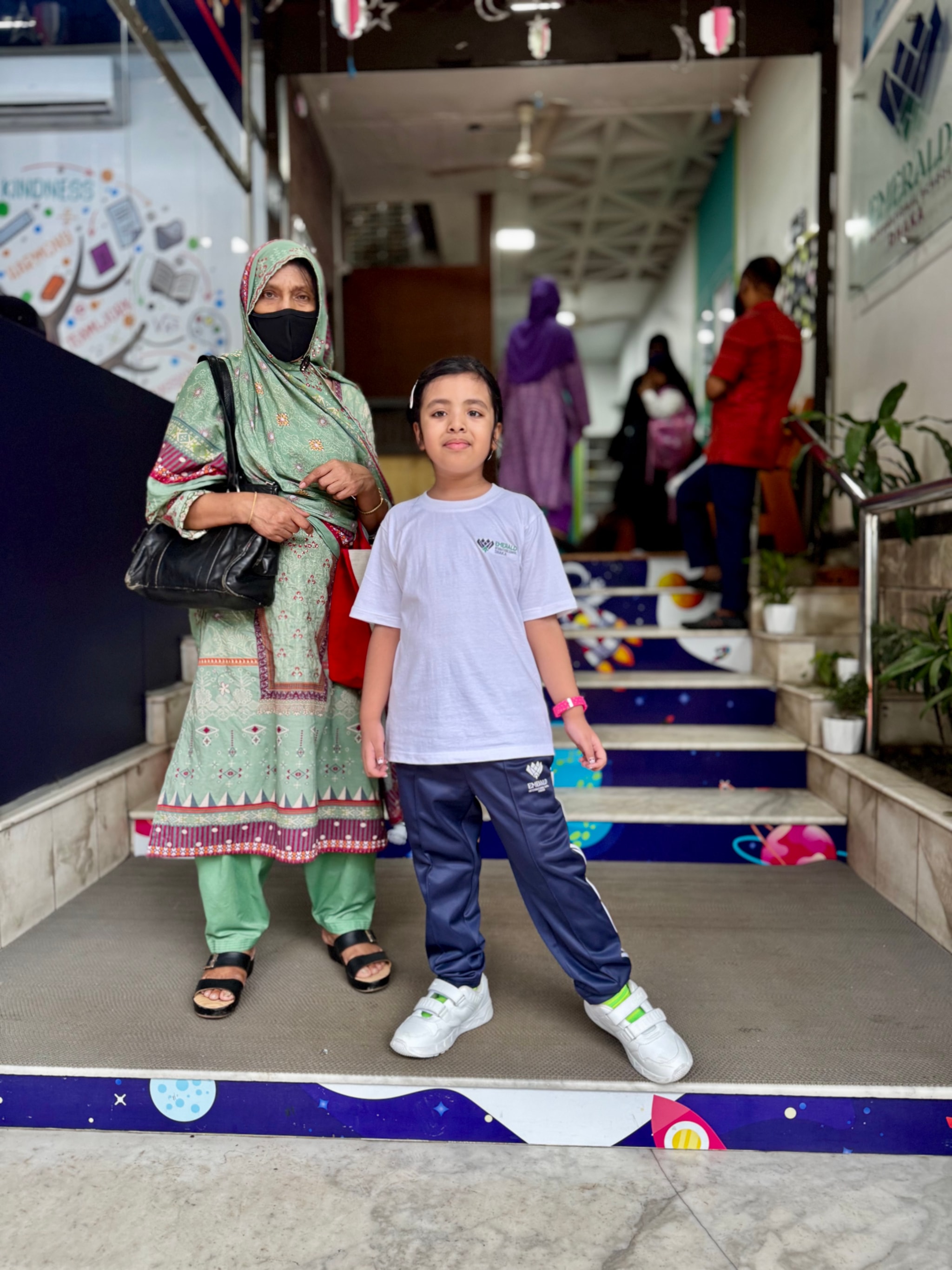 In the image, there are two individuals standing on a sidewalk. The person on the left appears to be an adult wearing a white shirt and black pants, with their hands in their pockets. They are also wearing a mask over their nose and mouth. On the right, there is a child dressed in casual attire, including a t-shirt with visible text that reads "SCOUT". Both individuals seem to be at a public outdoor location during daylight hours. In the background, there is a building with a sign that has text in Arabic. The sky above is clear, suggesting it might be early morning or late afternoon.