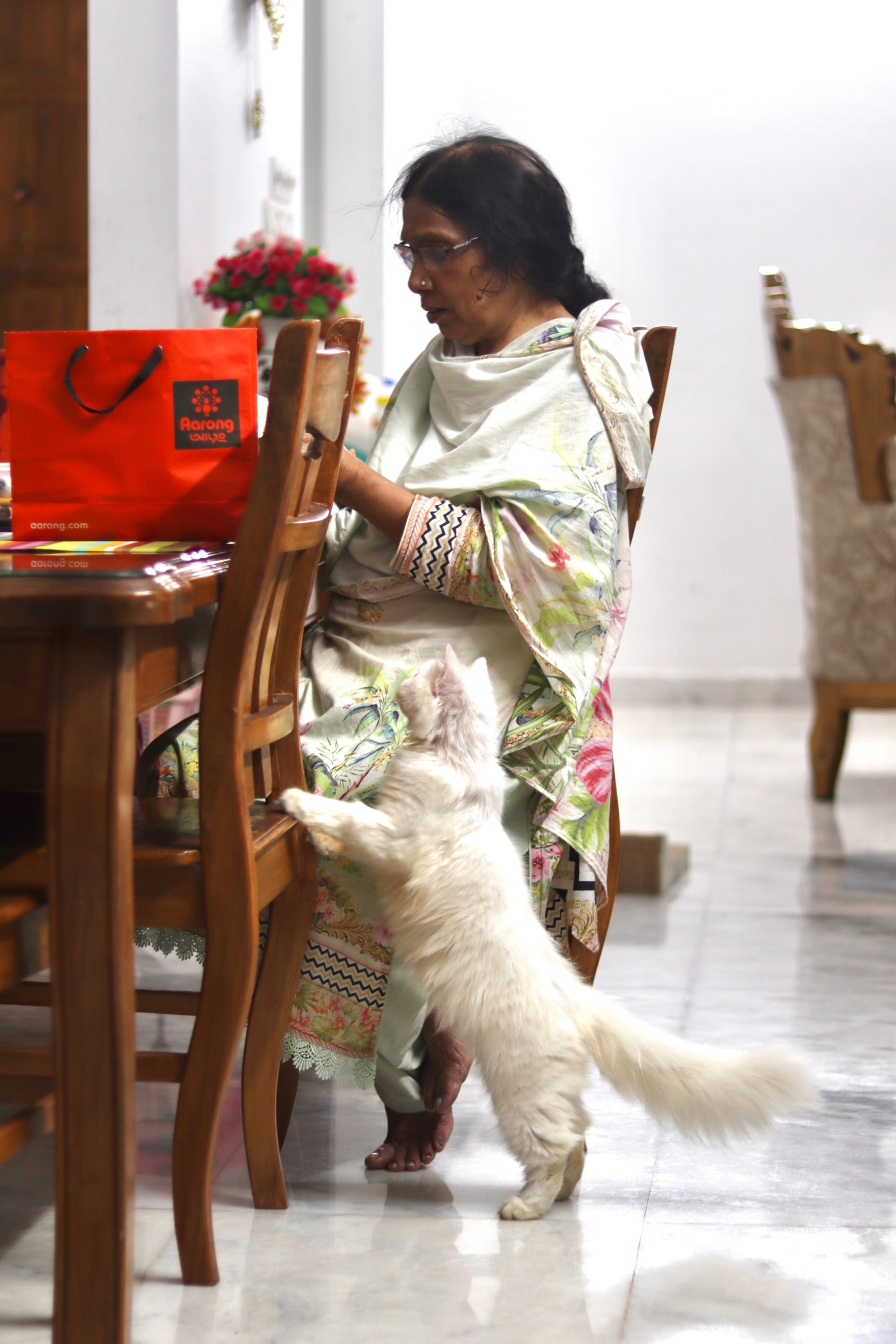 The image captures an indoor scene where a woman is seated at a table with a cat perched on the edge. She appears to be engaged in an activity, possibly using a cell phone which she holds in her hands. The woman has a traditional attire and is looking towards the camera. In front of her, there's a red shopping bag resting on the table. The setting seems to be a dining area with a chair visible next to the table. There are decorative elements like a potted plant in the background, and a flower vase is also present, adding to the homely ambiance.