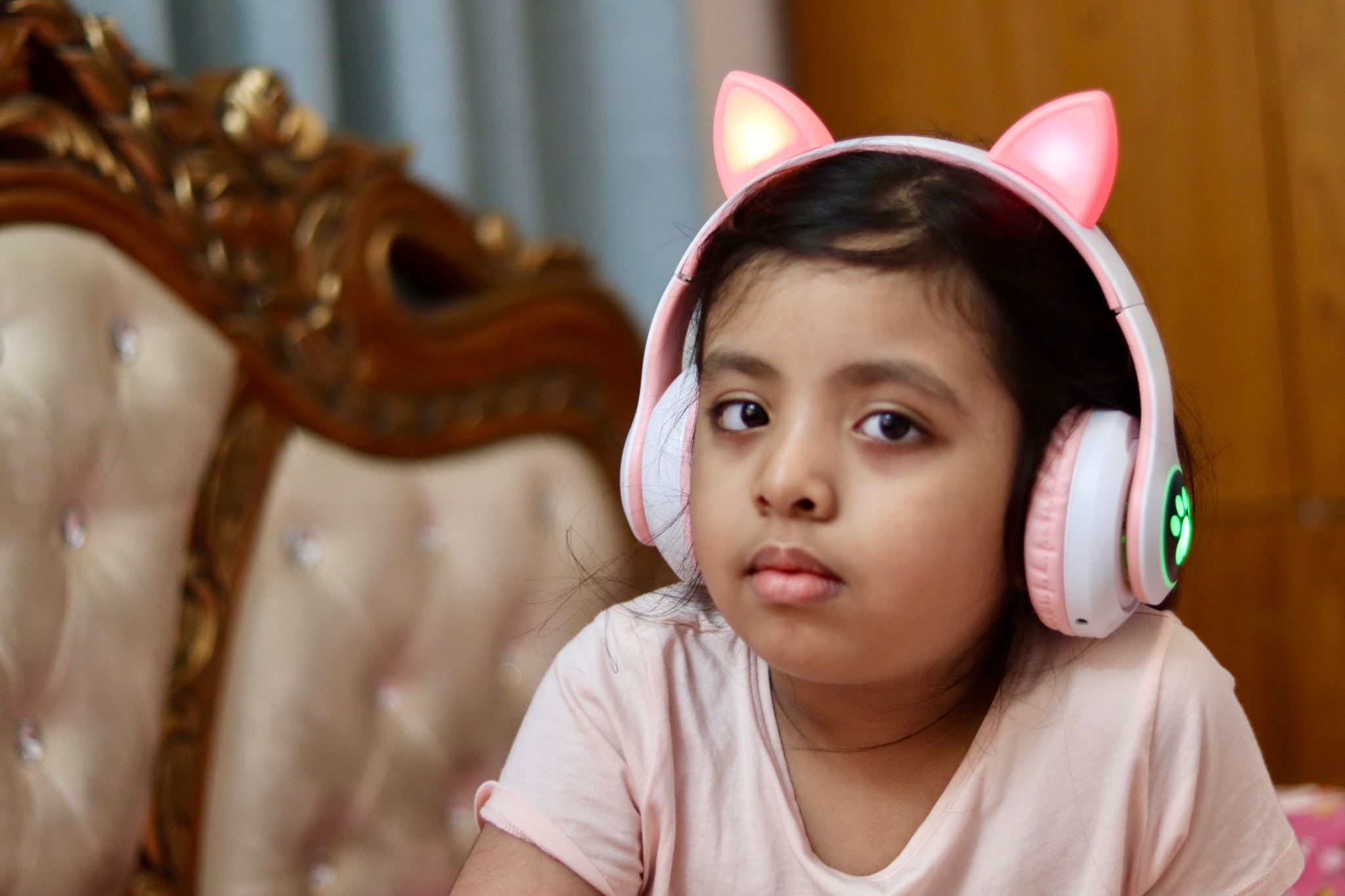 The image features a young child wearing headphones and looking towards the camera. The child has a gentle expression, possibly curious or attentive to something out of frame. They have pink cat ears on their head and are wearing a dark top with what appears to be white sleeves. There is no visible text within the image itself. Behind the child, there is a part of a decorative furniture piece with an ornate design on its backrest. The style of the photo seems candid, capturing a moment in the child's life rather than being posed for a specific occasion.