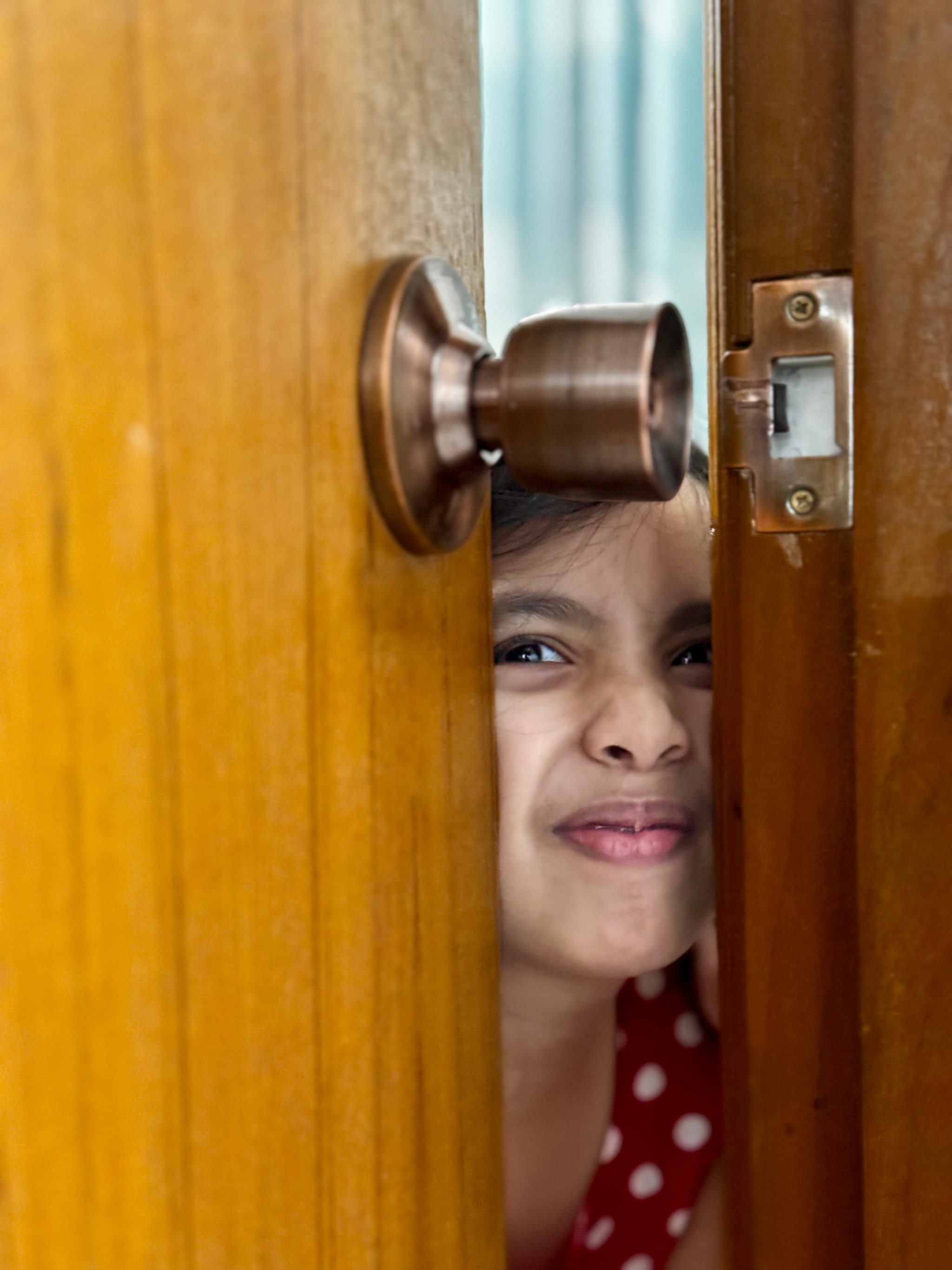 In the image, there's a person standing in front of a wooden door, looking through the door with a curious and surprised expression. The door has a brown finish and a bronze doorknob. The individual appears to be a young child or a young adult, dressed in casual clothing, peering out from behind the door, which is open a little bit at the top.
