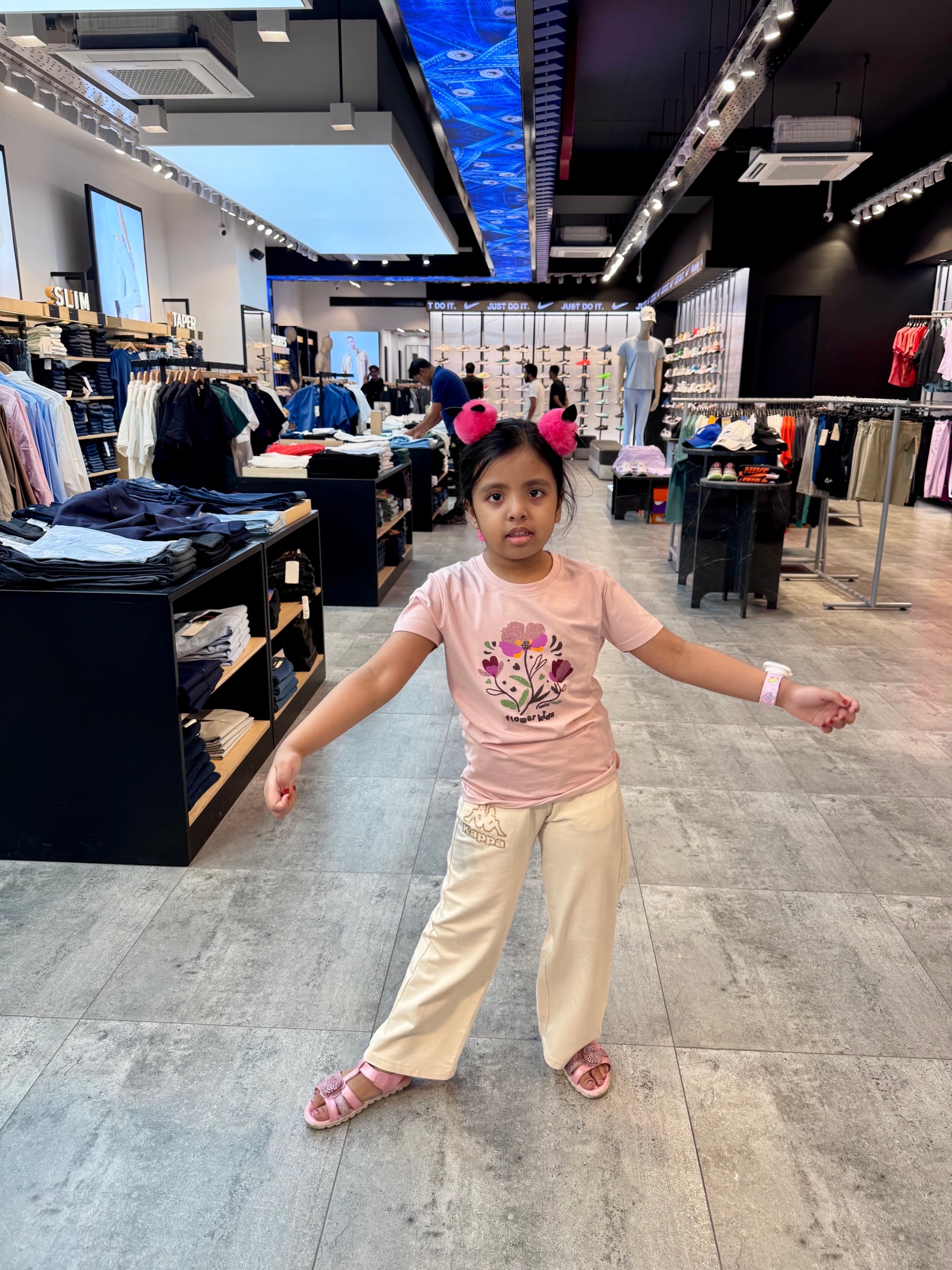 The image shows a young girl standing in an indoor retail space, possibly a store, with shelves and clothing racks in the background. She is wearing a pink top and white pants, with her arms outstretched as if she's dancing or posing for a photo. Her expression seems happy and energetic. There are various signs and logos visible on the storefront glass, including what appears to be a coat brand logo on the right side of the image. The lighting is artificial, suggesting an indoor setting with large windows or doors that allow natural light to enter.