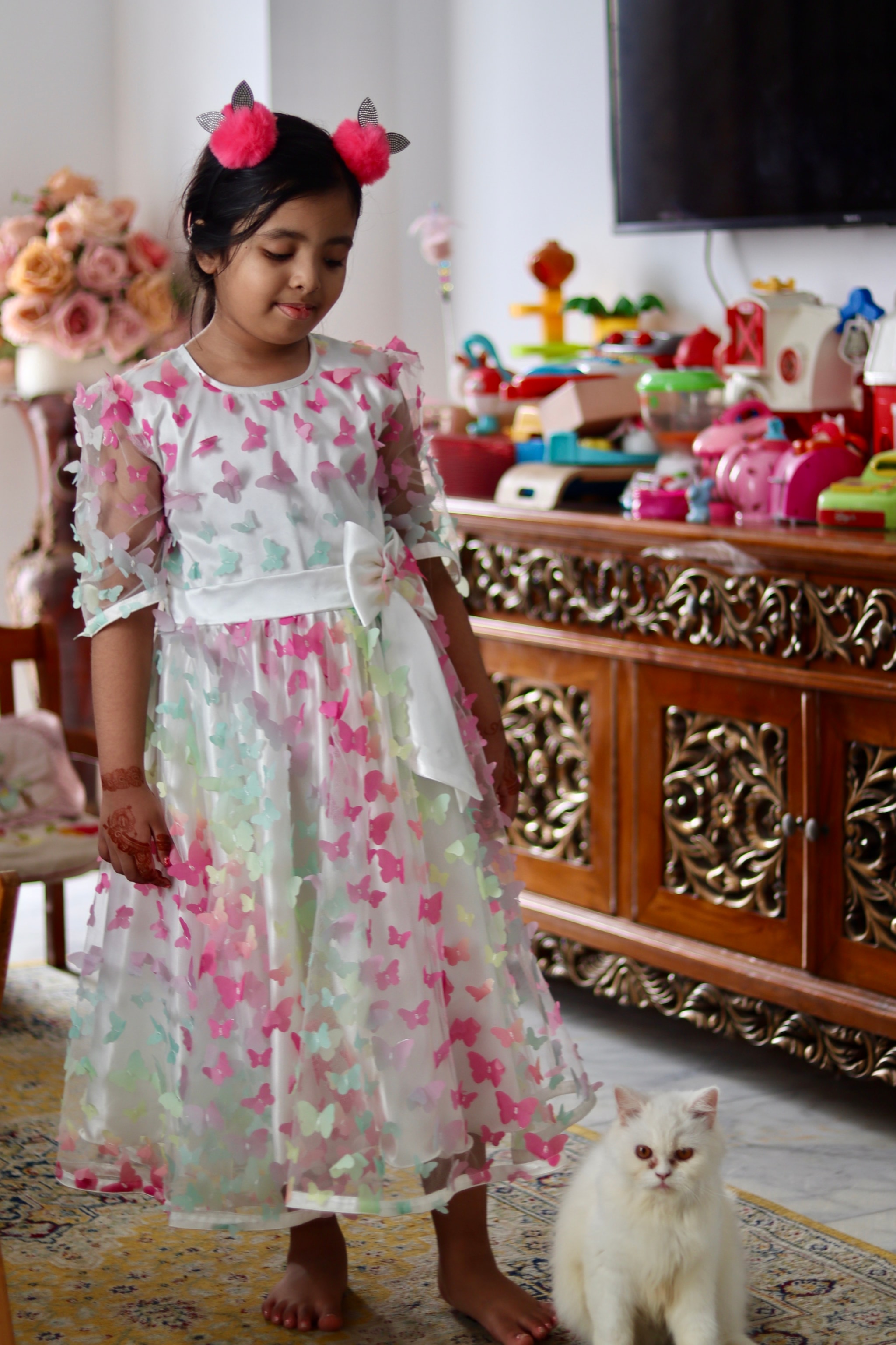 The image features a young girl standing indoors, looking towards a white and orange cat. She has dark hair, is wearing a colorful dress with a floral pattern, and has a pink bow in her hair. Behind her, there's a cabinet with various objects on it, including what appear to be toys or children's items. The style of the image suggests it may have been taken from a video frame due to the presence of a still image overlay, which is not part of the original photograph.