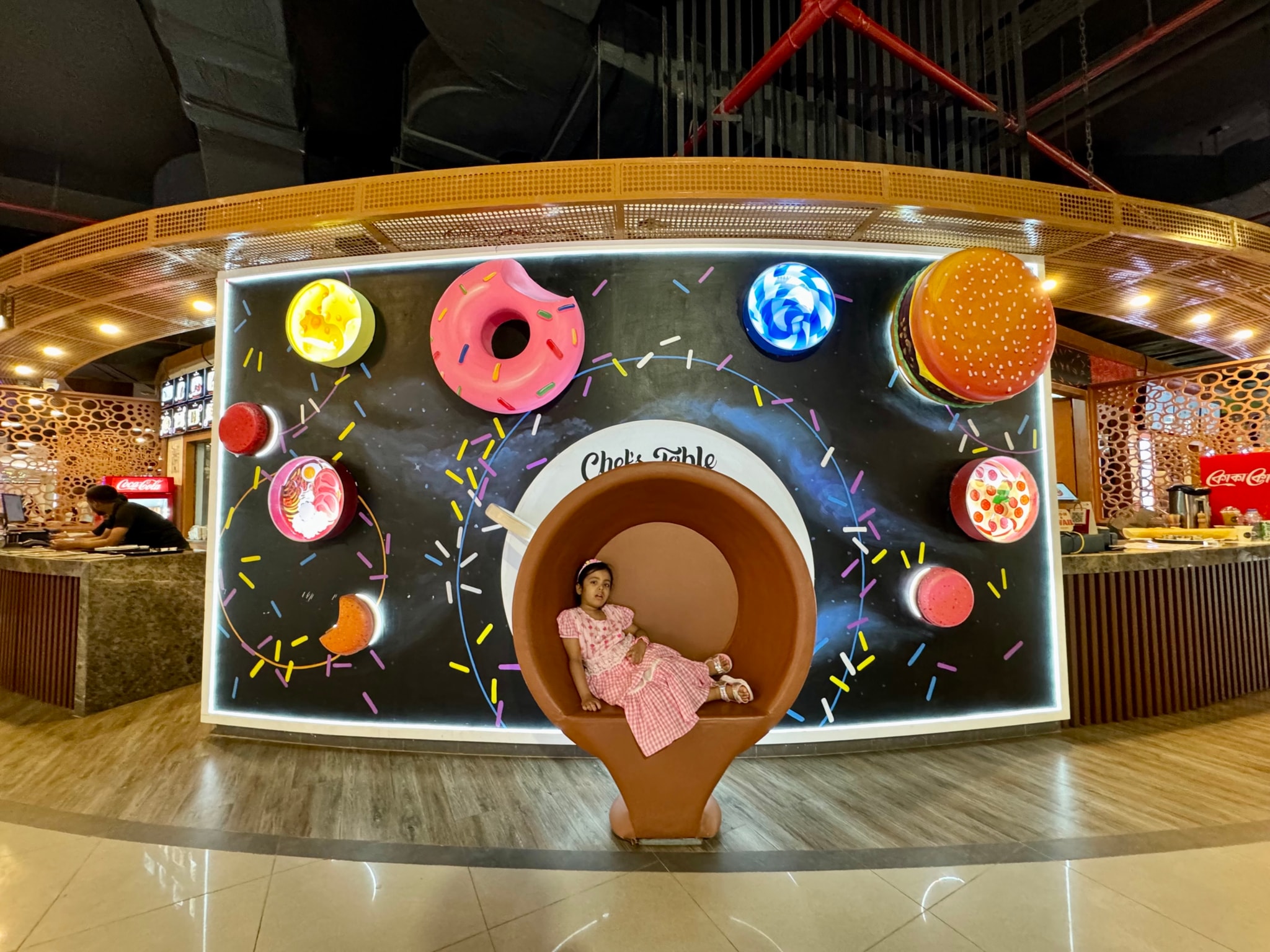 The image depicts an indoor setting with a playful and colorful design theme. There's an illusion of a woman sitting on a toilet paper roll, which is actually a part of the wall decoration featuring a creative arrangement of images and objects that mimic a real-life scenario. This artistic setup includes various items like donuts, planets, and other whimsical elements. The background shows a cafe or restaurant environment with a counter displaying pastries and beverages.