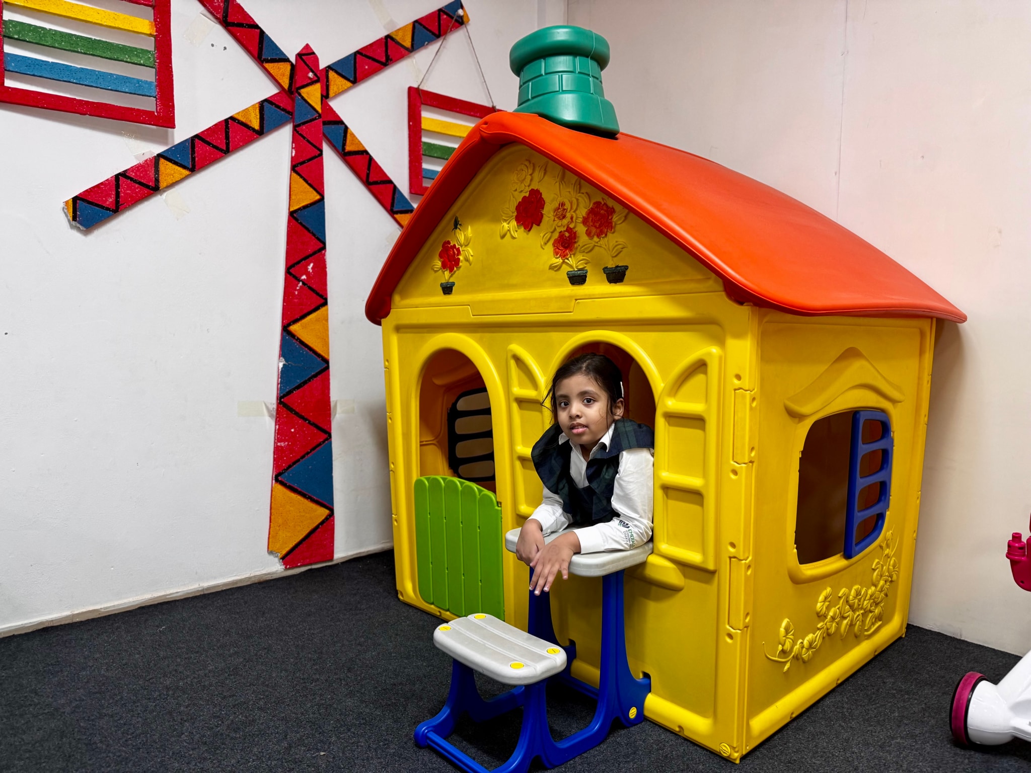 The image depicts an indoor setting where a child is sitting in a playhouse. The playhouse has a yellow roof and is shaped like a house, with a front door that's open. The child appears to be wearing a dark jacket and is seated on the floor inside the playhouse. There are colorful items hanging behind the child, including what looks like a fan with ribbons in hues of red, yellow, blue, and green. The environment suggests a casual indoor space that might be used for children's play or recreation activities.