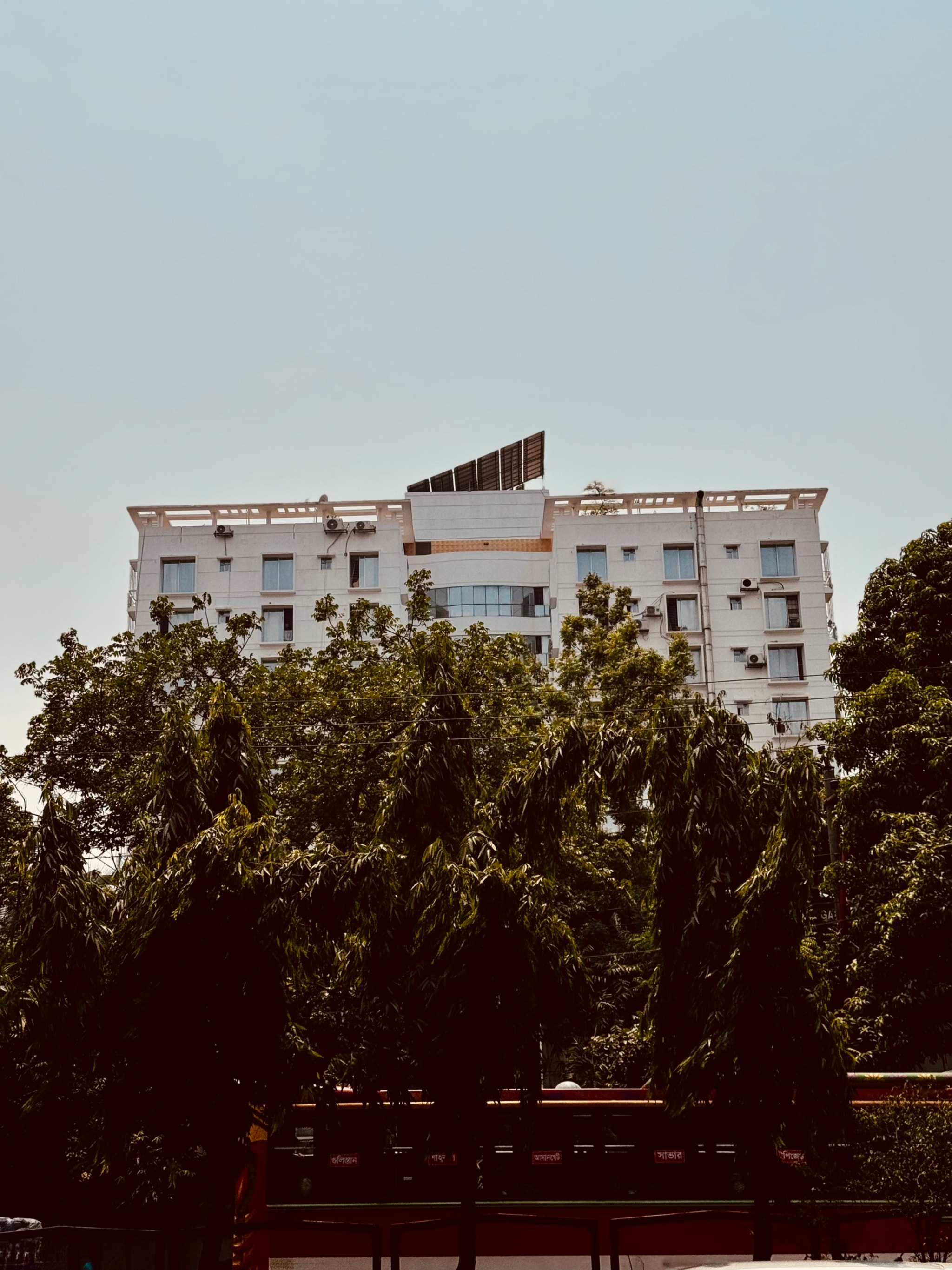The image displays a multi-story building with a visible balcony. The photograph has an overlay effect, causing parts of the structure to appear fragmented and distorted, creating a pixelated appearance. The background features a clear sky and lush greenery, which indicates that the building is surrounded by trees or plants. There's also a partial view of a power line. The style of the image appears to be digitally manipulated to create an artistic effect with the overlay.