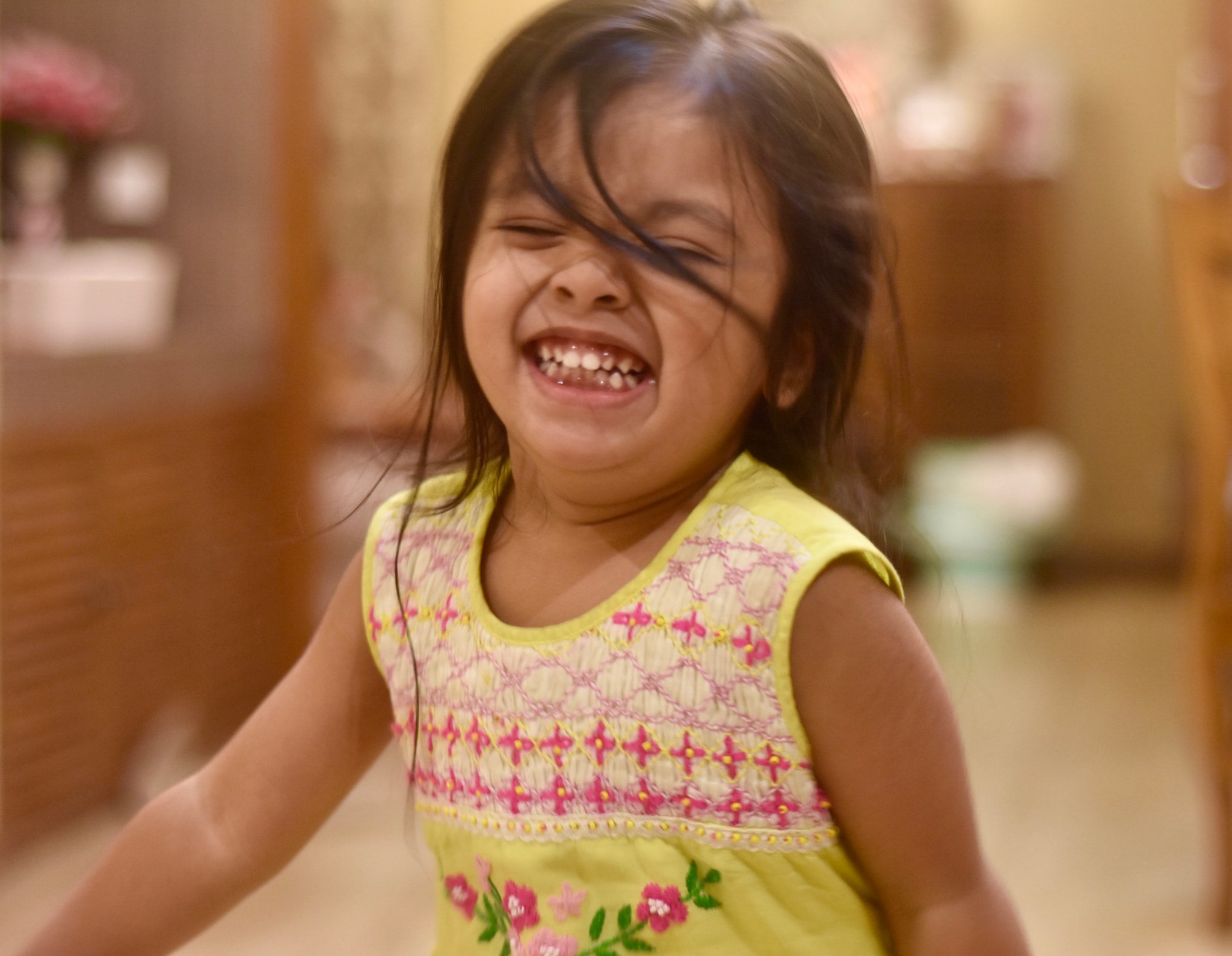 The image shows a young girl with her mouth open, laughing and displaying joy. She has dark hair and is wearing a light-colored sleeveless top with a pattern on it. The background is blurred but appears to be an indoor setting with a cabinet and a counter visible.