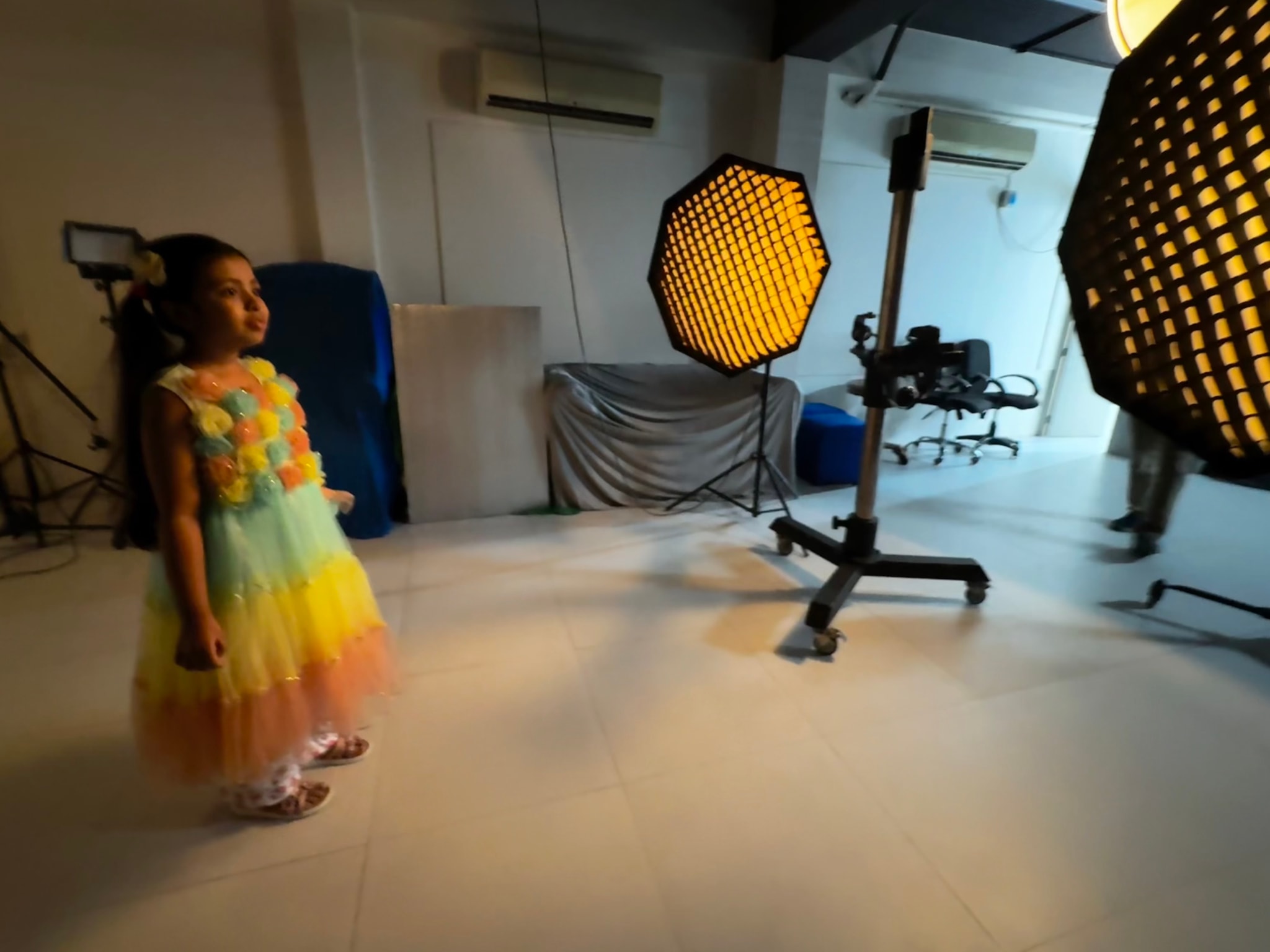 This image features a child standing in what appears to be a photography studio. The child is looking at the camera, wearing a colorful outfit, with their arms by their sides. In front of them on the floor are several items that resemble photo backdrops or props, including what seems like a honeycomb pattern and possibly other elements for photoshoots. The background is dimly lit, which suggests it might be evening or in a room without artificial lighting. The setting gives an impression of being in a professional photographic environment.