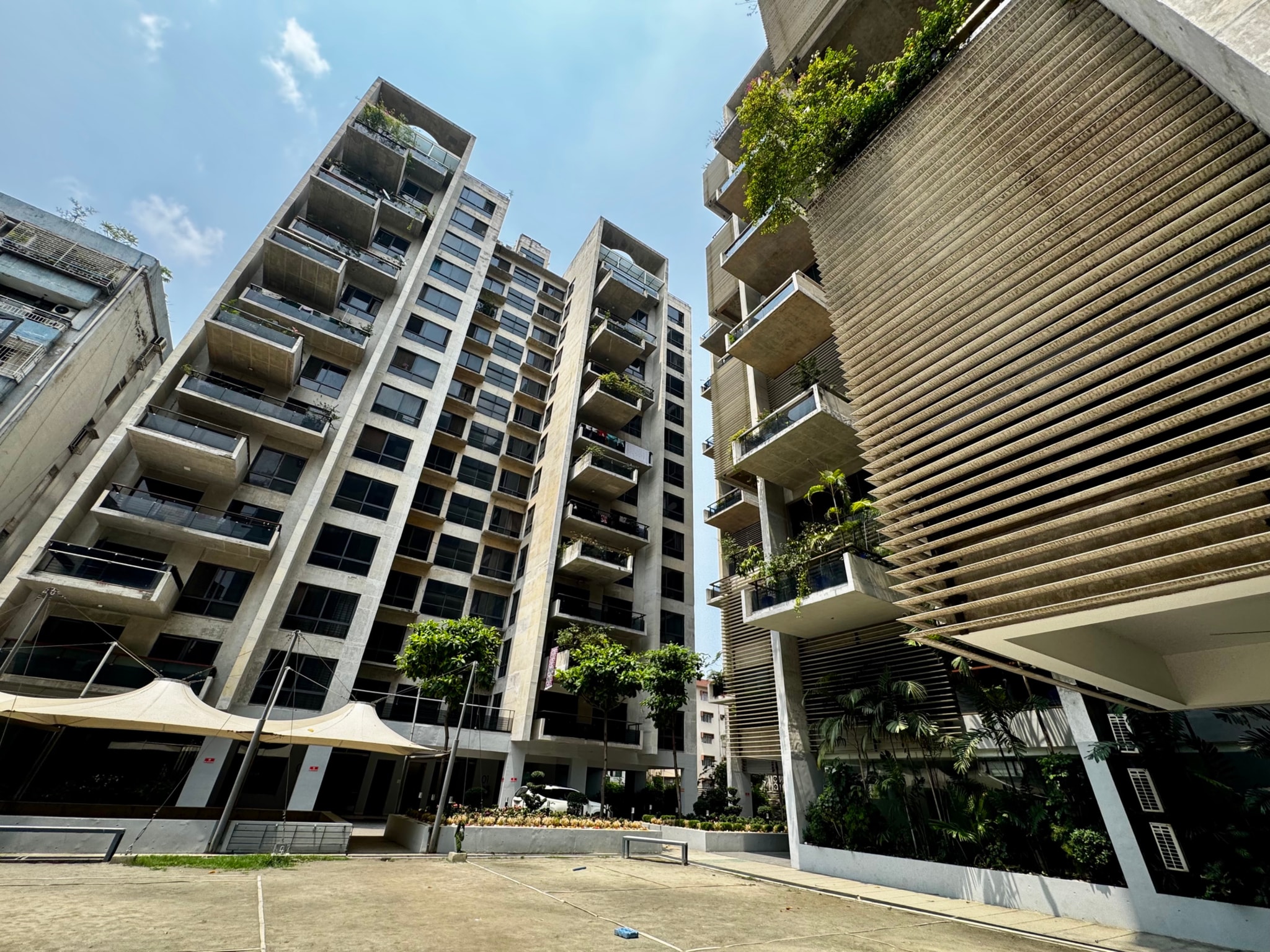 The image depicts an urban residential area. It features modern, multi-story buildings with balconies and exterior design elements such as vertical gardens on some balconies. The architecture suggests contemporary design with sleek lines and glass facades. The sky is clear with minimal clouds, indicating fair weather conditions.