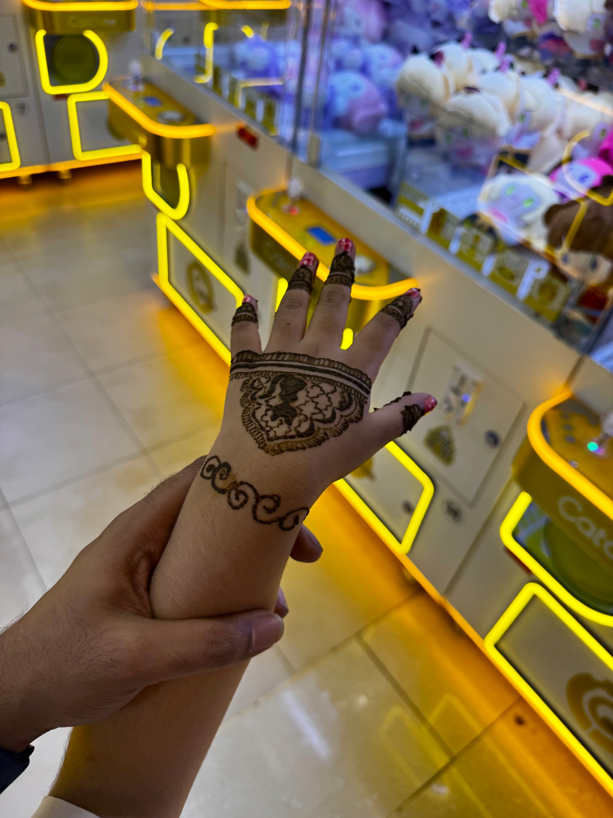 The image features a person's hand with a henna-like tattoo on their finger, reaching towards the camera. In the background, there's a display case with various items inside, and another display area with shelves full of merchandise. There are colorful lights illuminating the scene, suggesting that this could be an indoor space such as a market or exhibition.