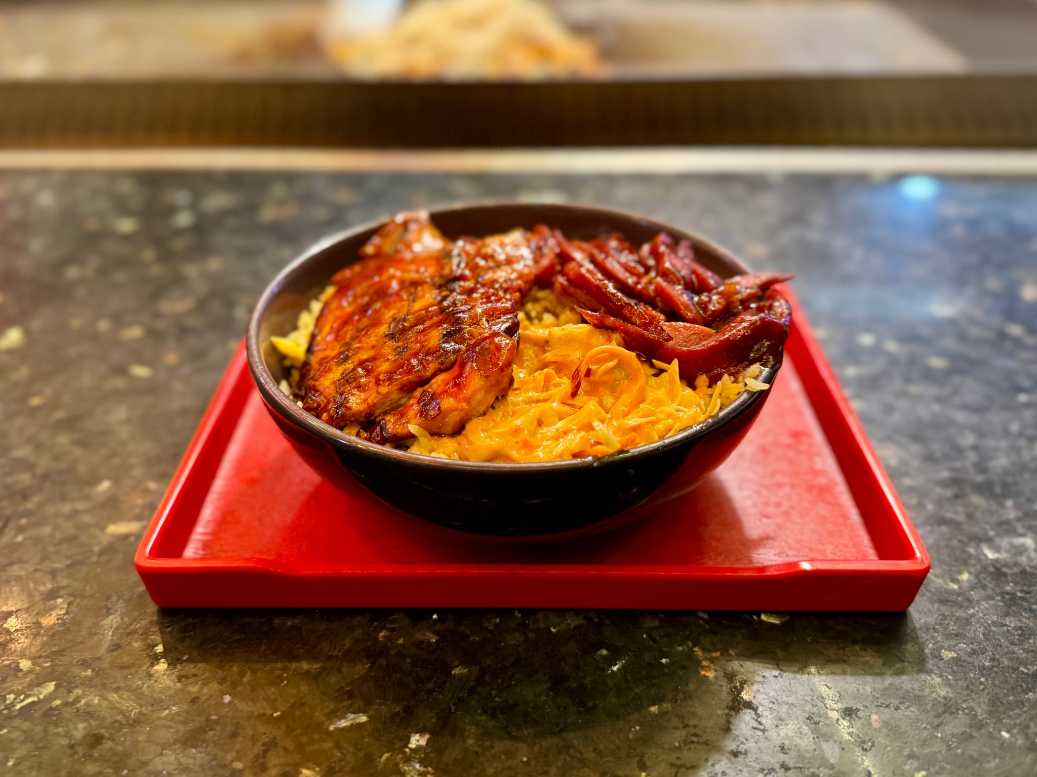 The image features an oriental meal served on a red tray atop a countertop. There are two bowls: one with what appears to be steamed rice and another with slices of grilled meat, possibly chicken. Beside the dishes are a small bowl filled with a sauce that could be chili or curry, and a portion of fried noodles in a separate bowl. The tabletop has a dark surface, and the overall setting suggests a casual dining environment. There is also a blurred image of what looks like another food item in the background, possibly part of the same meal or from a nearby station.