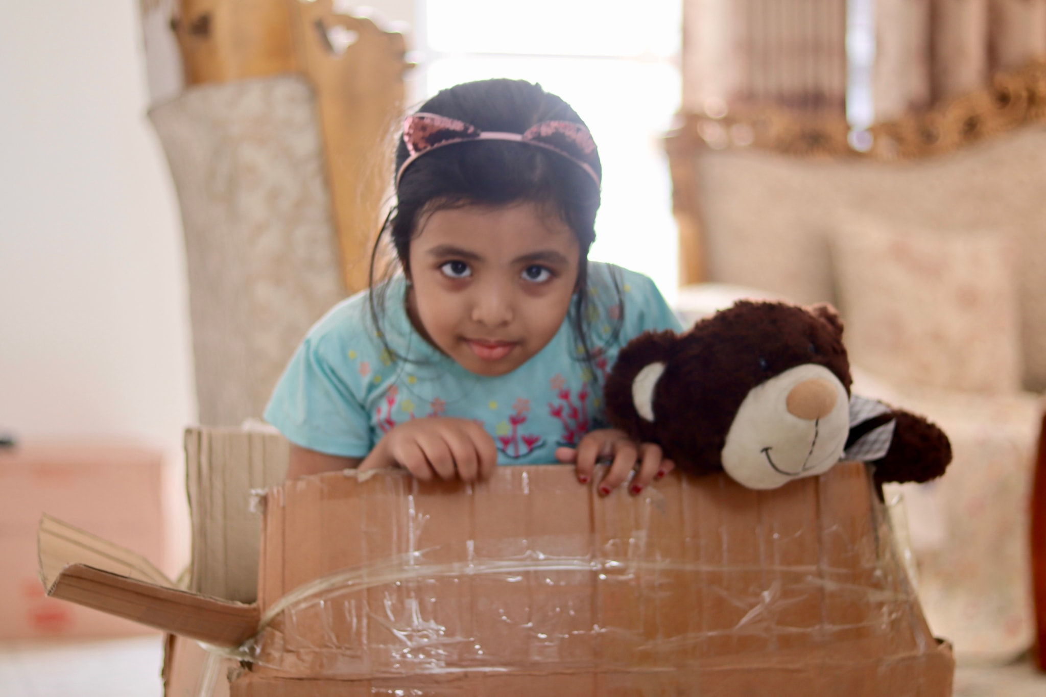 The image depicts a young girl sitting on top of what appears to be a cardboard box. She's looking directly at the camera with a playful expression. There's a teddy bear next to her, and it seems like she might be in a room undergoing some sort of construction or renovation work, as suggested by the visible walls and materials. The girl appears to be enjoying her time, perhaps playing or exploring.