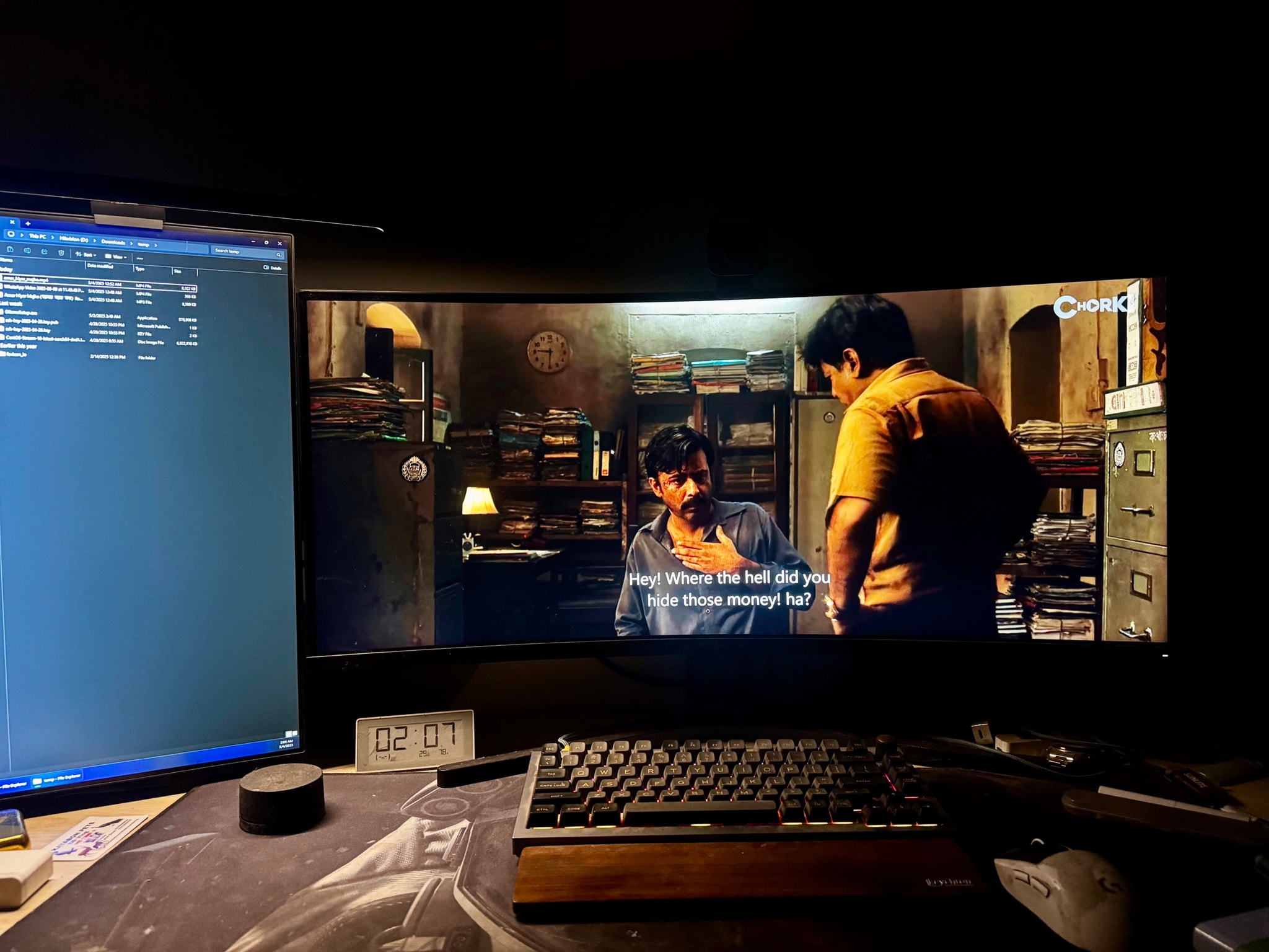 The image depicts a home theater setup with a large widescreen monitor prominently displaying a movie scene. Two individuals are visible in the scene, engaged in what appears to be an intense conversation. There's a well-lit room, with a gaming computer partially visible on the left side of the frame.