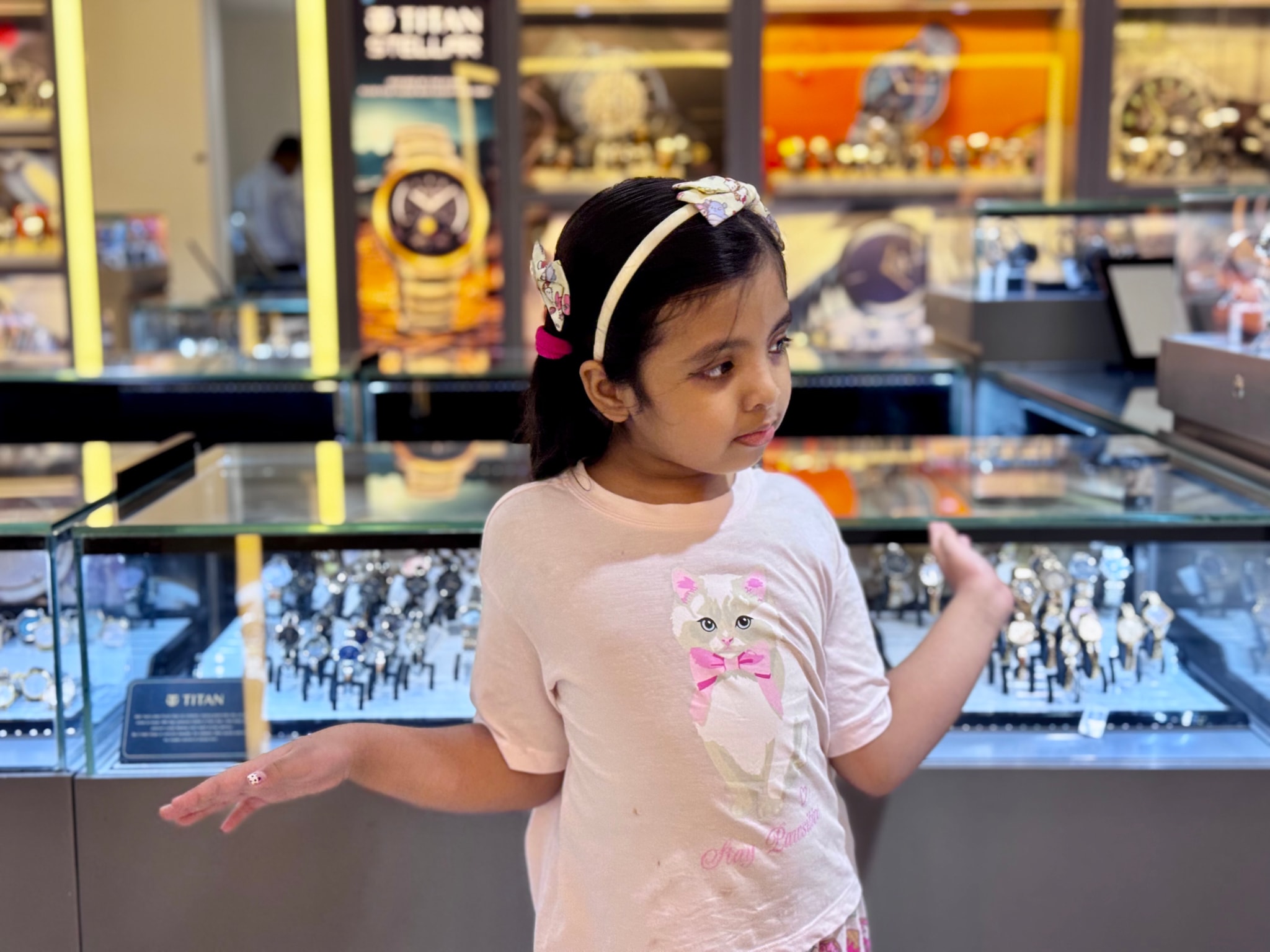 The image depicts a young girl with light hair, wearing a headband and a dress with floral patterns. She stands in front of a display case containing various jewelry items wrist watches. In the background, there are store displays with product signs and merchandise. The lighting suggests an indoor setting, likely a jewelry store or exhibition.
