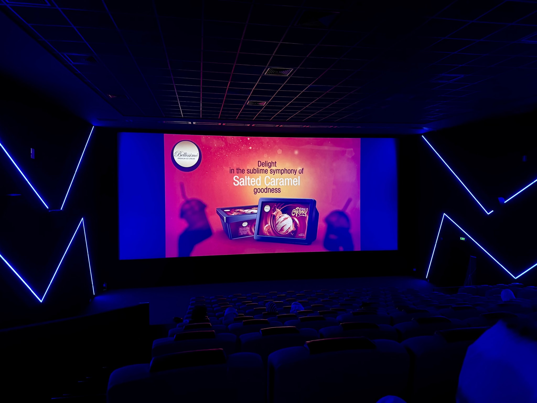 The image appears to be taken inside an auditorium during a movie screening. There's a blue light on the walls and a screen in the background displaying what seems to be Indian film content. The focus is slightly blurred, but you can discern that there are people sitting in the audience.