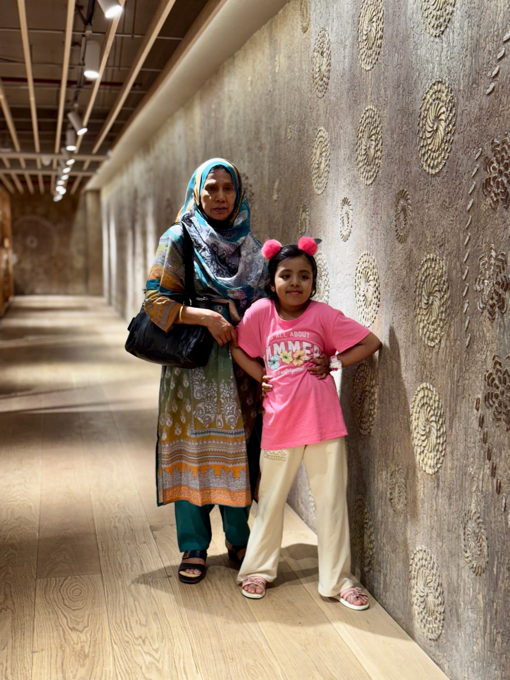 The image depicts an indoor setting, possibly a corridor or hallway with walls adorned with what appears to be decorative elements. There are two individuals present in the scene; an adult and a younger child, both looking towards the camera. They seem to be standing close to each other. The adult is wearing traditional South Asian attire, while the child has on casual clothing including a pink t-shirt. Both of them have handbags with them. The lighting in the room is even, and the floor appears to be made of polished wood or stone.